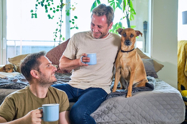 Gay couple of two mature guys on the sofa at home with a cup in hand and their dog next to them