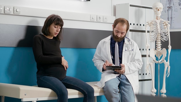 General practitioner doing checkup examination with patient expecting baby, giving medical advice about childbirth. Medic talking to woman with pregnancy belly at consultation.