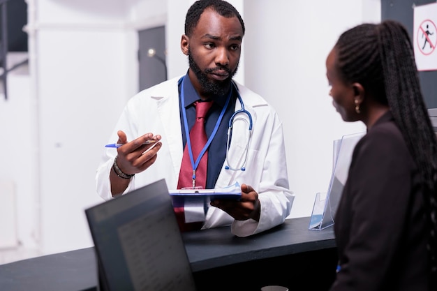 General practitioner talking to receptionist about healthcare insurance support and appointments at reception counter. Physician discussing about checkup visits with medical worker in clinic lobby.