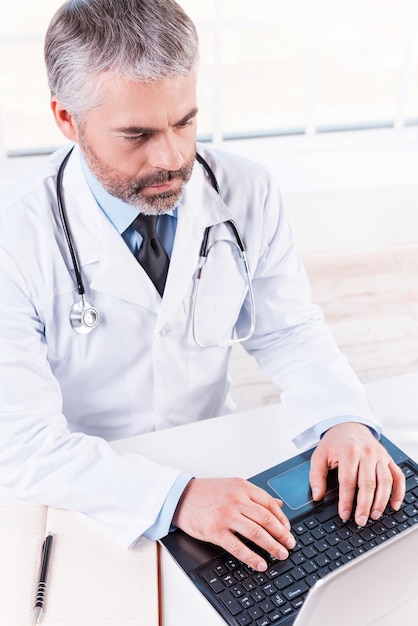 General practitioner at work. Top view of confident mature grey hair doctor working on laptop while sitting at his working place