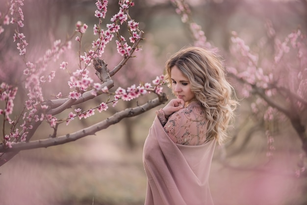 Gentle portrait young curly blonde woman in brown pleated skirt pink blouse covered shoulders with stole stands in blooming peach gardens at sunset