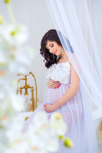 Gentle pregnancy. Beautiful pregnant in light white dress with orchids.