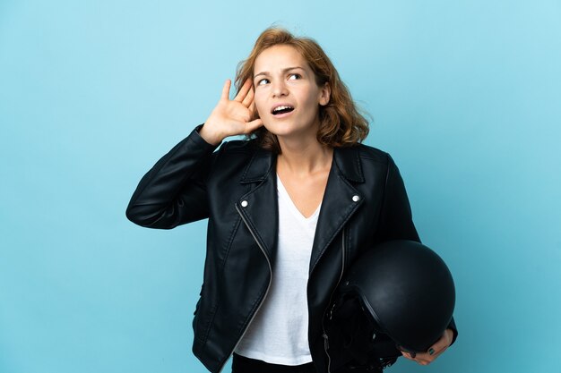 Georgian girl holding a motorcycle helmet isolated on blue wall listening to something by putting hand on the ear