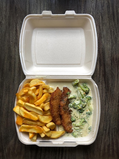 german takeaway food: schnitzel (breaded escalope) with french fries and broccoli cheese sauce in polystyrene container on rustic wooden table