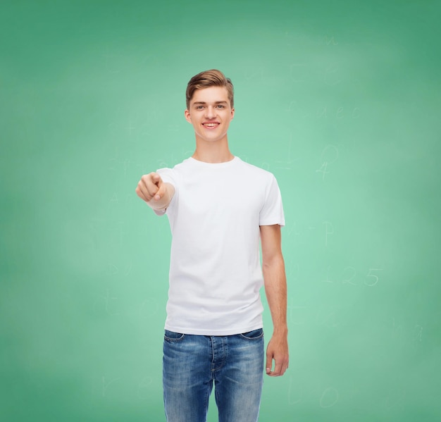 gesture, advertising, education, school and people concept - smiling young man in blank white t-shirt pointing at you over green board background