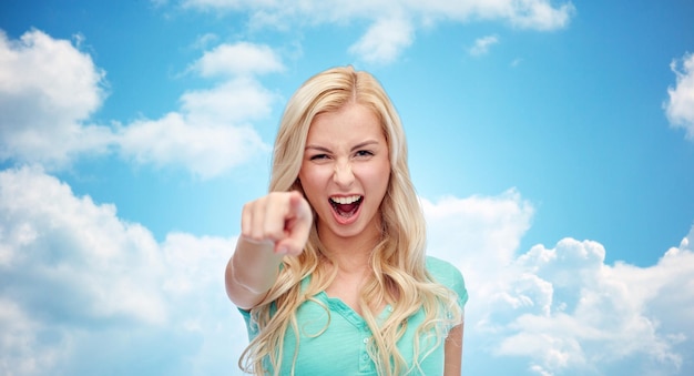 gesture, emotions, expressions and people concept - happy smiling young woman or teenage girl pointing finger to you over blue sky and clouds background