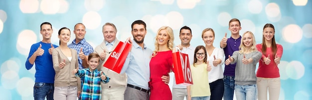 gesture, sale, shopping and people concept - group of smiling men, women and boy showing thumbs up over blue holidays lights background