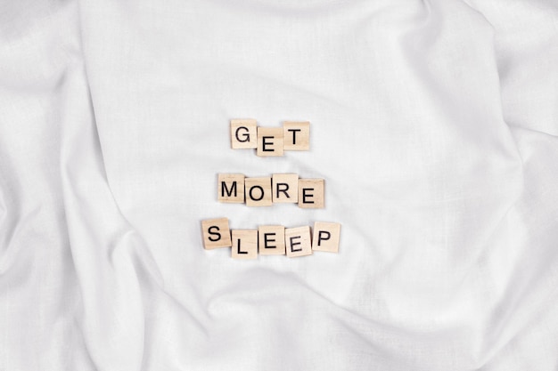 Get more sleep phrase made of wooden letters flat lay on white fabric background top view