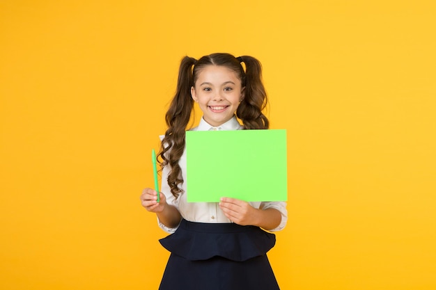 Get a writing pen Little girl holding blank paper sheet for writing task on yellow background Small child display green poster for writing information Empty show card for writing copy space