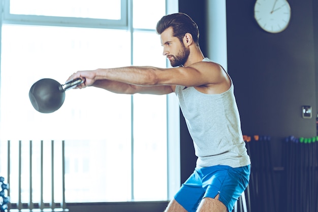 Getting stronger every day. Side view of young handsome man in sportswear working out with kettle bell at gym