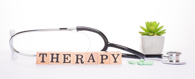 Getting therapy concept. Horizontal panoramic closeup view photo image of doctor stethoscope succulent pile of green capsules and wooden blocks making word therapy isolated white backdrop