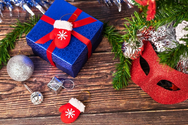 A gift in a blue wrapper, tied with a red ribbon, stands on a wooden table, with silver tinsel, Christmas balls and small decorative red mittens, a masquerade mask.