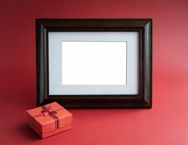 Gift box and photo frame Empty white blank rectangle space in old vintage wooden photo frame with small red present box on red background Template for photo or card with a gift minimal style