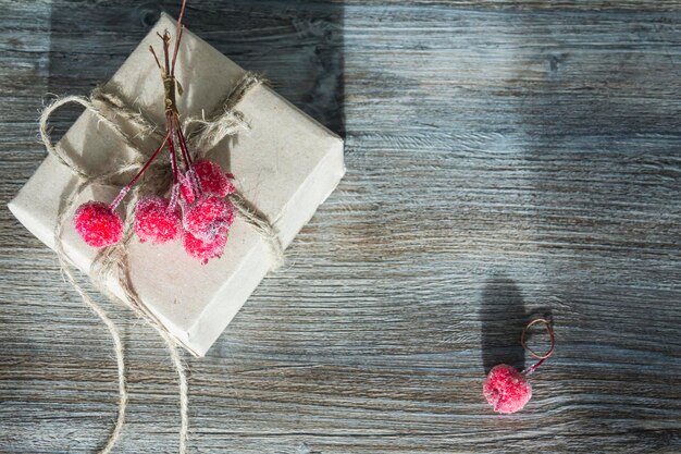 A gift box with red berries packed in kraft paper and tied with linen twine on a wooden background