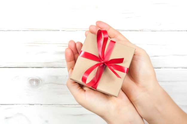 Gift box with red ribbon and bow in giving hands on white wooden