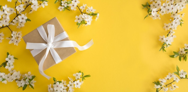 Gift box with tied white bow and flowering tree branches on the yellow backgroundTop view Copy space