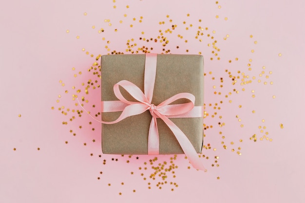 Gift box wrapped in craft paper with pink ribbon on pale pink with glitters. Festive concept.