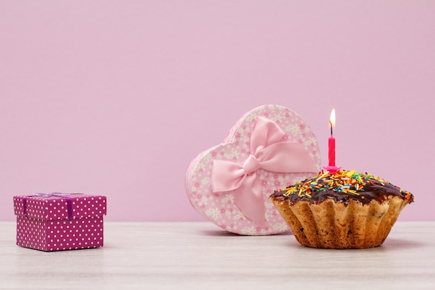 Gift boxes and tasty birthday muffin with chocolate glaze and caramel, decorated with burning festive candle on lilac background. Happy birthday minimal concept.