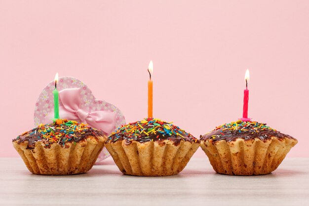 Gift boxes and tasty birthday muffins with chocolate glaze and caramel, decorated with burning festive candles on wooden and pink background. Happy birthday concept.