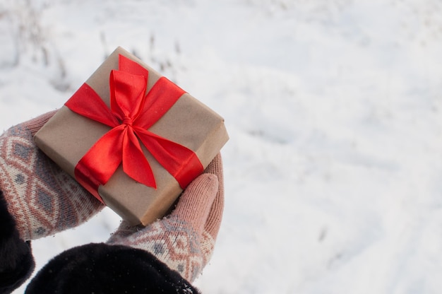 A gift in craft paper and with a red ribbon in the hands of gloves on a winter blurry background