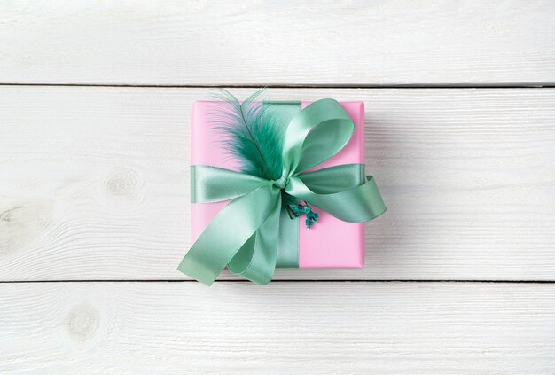 A gift in a pink package with a mint ribbon and a feather on a white wooden background.