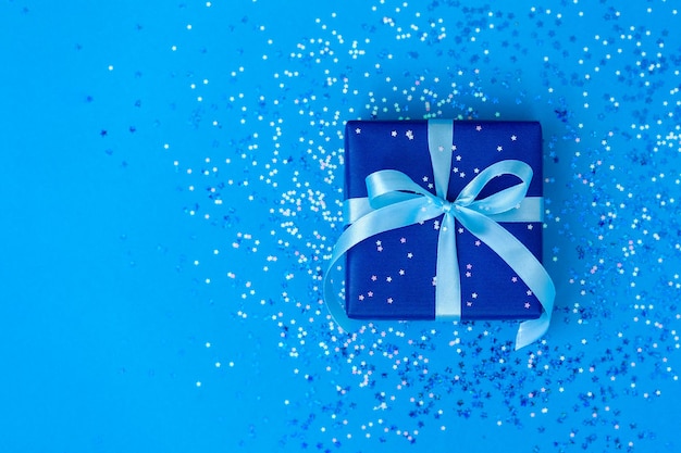 Gift or present box tied with a blue ribbon on blue paper background with sparkle