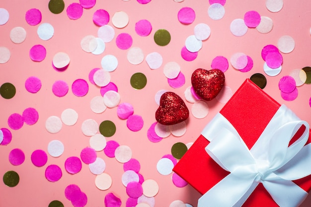 Gift and red shiny hearts on a pink table. Flat lay, top view.