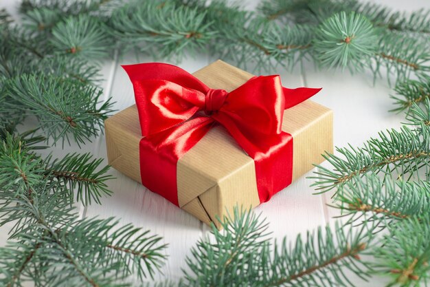 Gift wrapped in craft paper and red ribbon with green branches of a Christmas tree on a white wooden background.