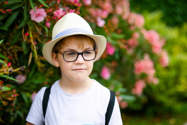 Ginger boy in the straw hat and big glasses near the green bush with pink flowers in the summer park