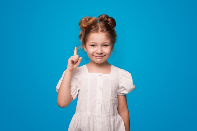 Ginger girl pointing up with forefinger wearing a white dress on a blue  wall