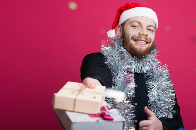 Ginger man with beard and santa hat is giving at front a present smiling on a red  wall