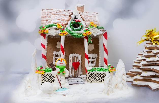 Gingerbread house, gingerbread Christmas tree and a sugar mastic snowman on background of defocused silver lights. Mockup for seasonal offers and holiday post card