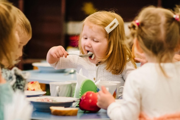 Photo girl 3 years sits at the table and independently eating kids eat in kindergarten lunch