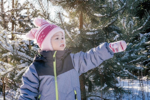 Girl 34 years old in winter overalls hat and mittens stands against snowcovered pines and fir looks away pointing