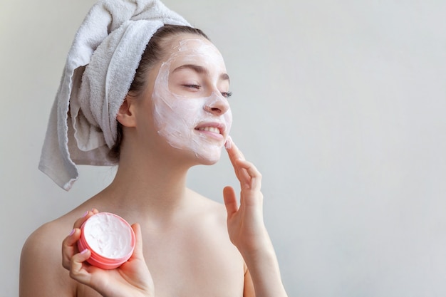 Girl applying white facial mask isolated on white background. Young woman in towel on head with white nourishing mask or creme on face. Skin care treatment spa natural beauty and cosmetology concept.