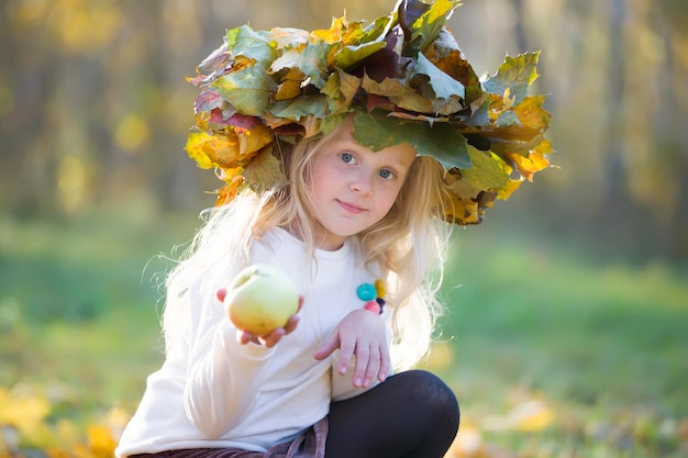 Girl in autumn park with a wreath of yellow leaves with an apple A child on a walk in the fall