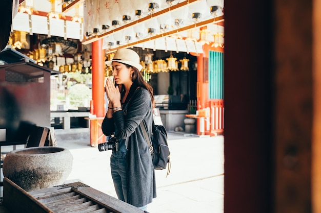 girl backpacker experience japanese culture in shitennoji. young lady tourist clap her hands closing her eyes and quietly praying in the peaceful temple. white lanterns hanging up on the roof.