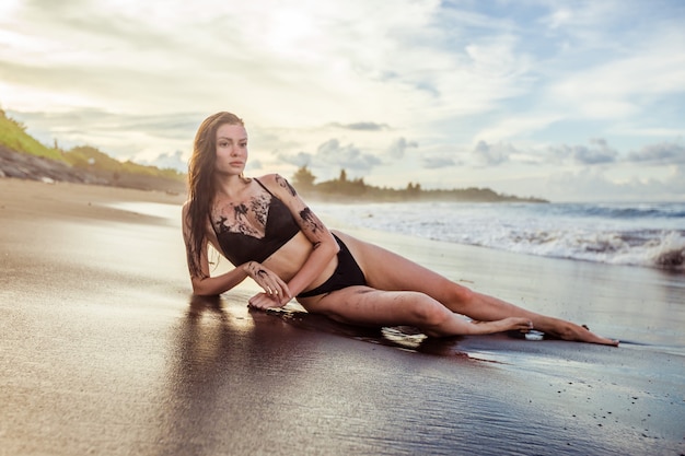 The girl in a bathing suit covered with black sand is lying on the beach