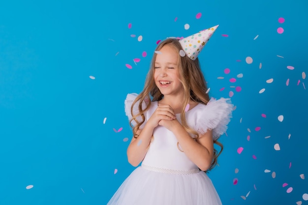 girl in a beautiful dress and a cardboard hat stands on a blue background and throws confetti