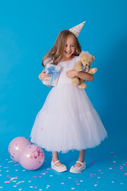 girl in a beautiful fluffy white dress holds a teddy bear and a gift box in her hands