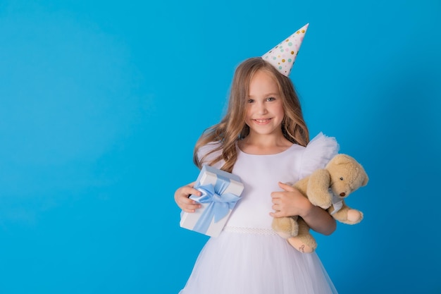 girl in a beautiful fluffy white dress holds a teddy bear and a gift box in her hands