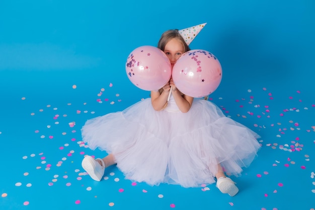 girl in a beautiful white fluffy dress and a festive hat is sitting on the floor on blue background