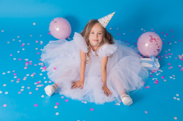 girl in a beautiful white fluffy dress and a festive hat is sitting on the floor on blue background