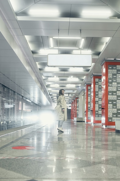 Girl in a beige trench coat in the subway waiting for the train on the platform.