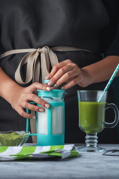 A girl in a black apron is preparing a latte from matcha green tea.