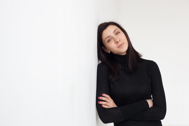 A girl in black clothes is standing by the white wall She looks confidently forward arms crossed by her chest