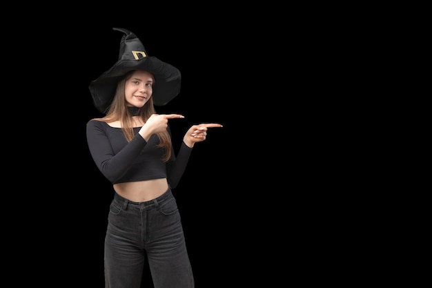 Girl in black clothes and pointed hat pointing her index finger sideways on empty space for your text Black background