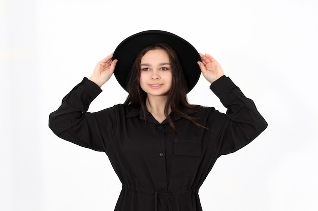 A girl in a black hat poses and smiles on a white background holding her hat