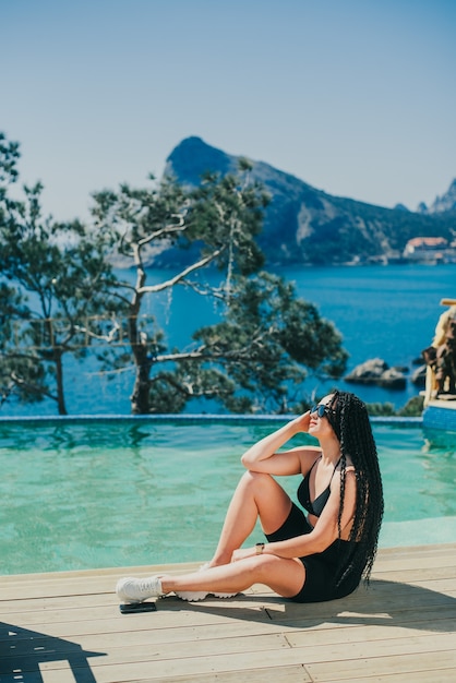 A girl in black sitting by the pool on a beautiful background of the sea and mountains, with braided afro-curls on her head