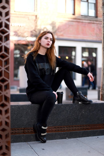 Girl in black stylish clothes sits outdoors with coffee. City Style. Red-haired model.